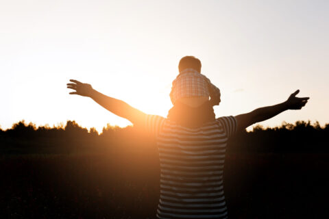 Father and son silhouette walking on the field at the sunset time, boy sitting on mans shoulders. Concept of happy family weekend